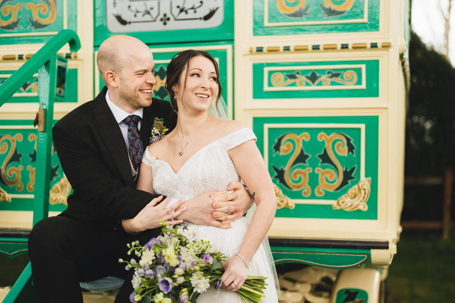 A couple sit on the steps of a green Romany Caravan at South Farm. The caravan is intricately decorated with gold flourishes.
