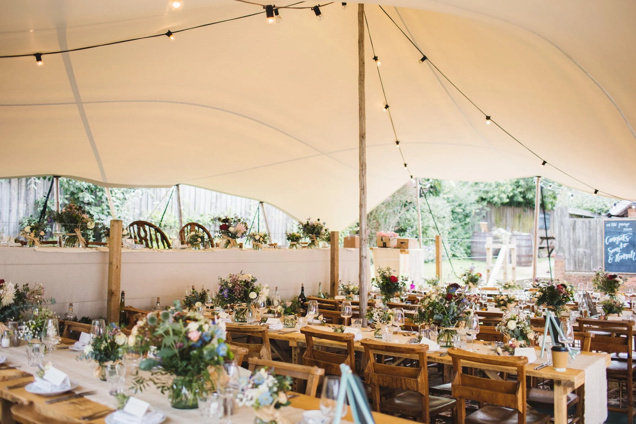 An interior view of the stretch tent in the garden at The Sun in Felmersham. Festoon lights hang from the ceiling over wooden tables covered in wildflower arrangements.