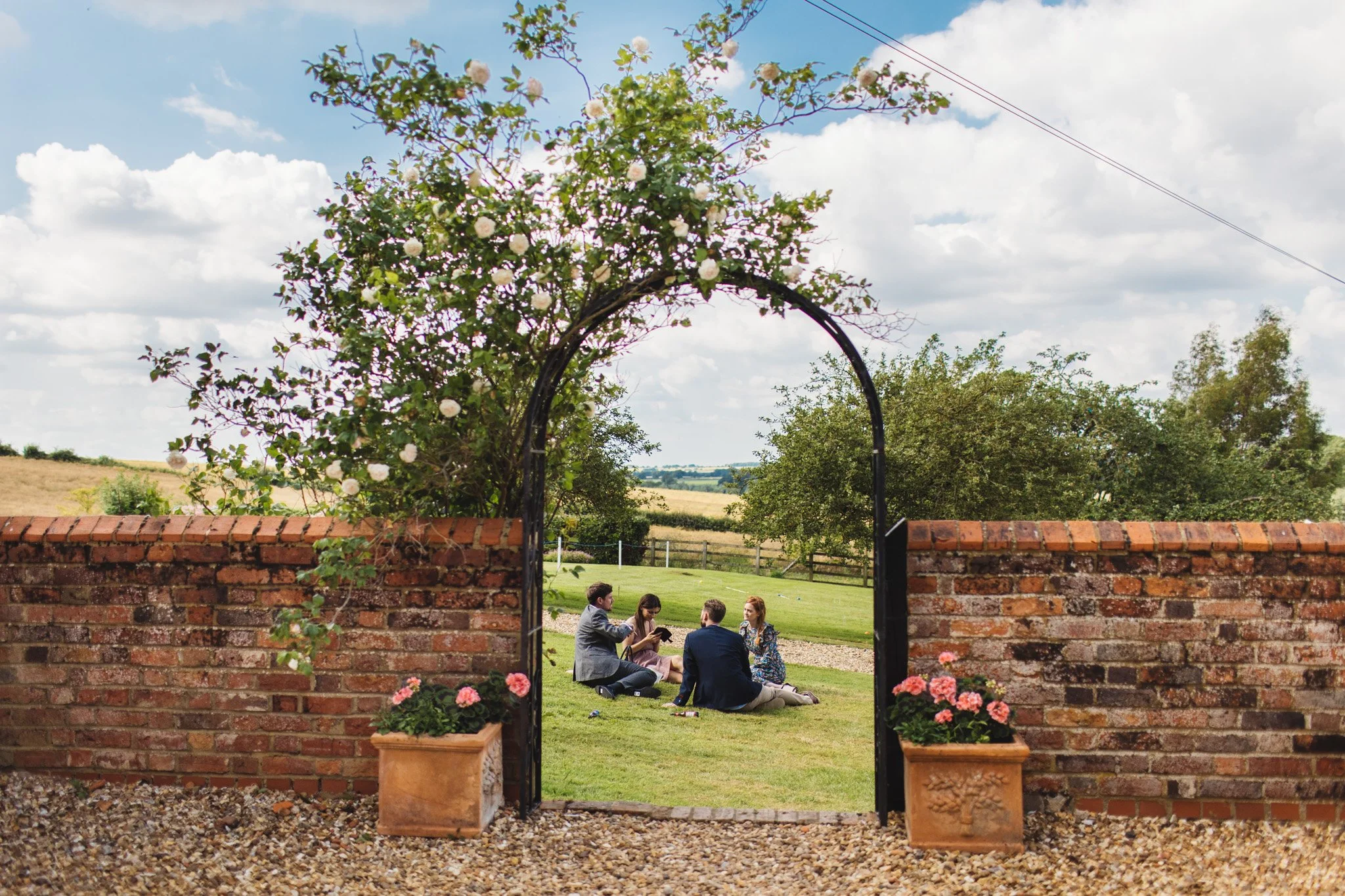 Guests sit on the lawn, relaxing with a drink. They are framed by a rose arch and blue sky.