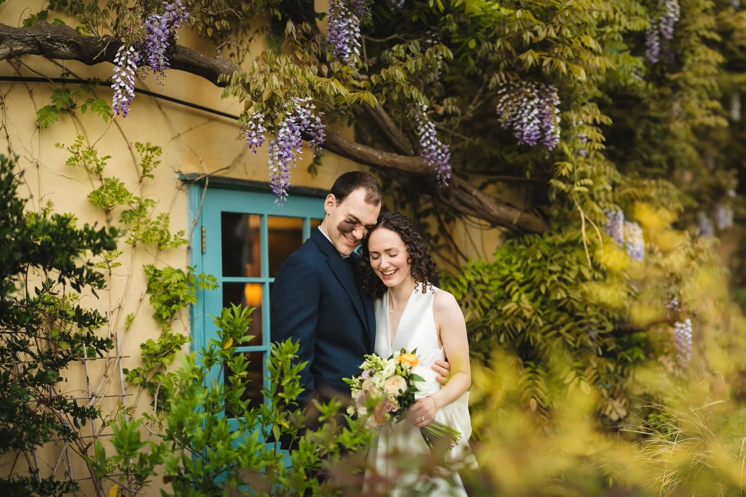 A bride and groom cuddle up in the South Farm gardens. Purple wisteria surrounds them.