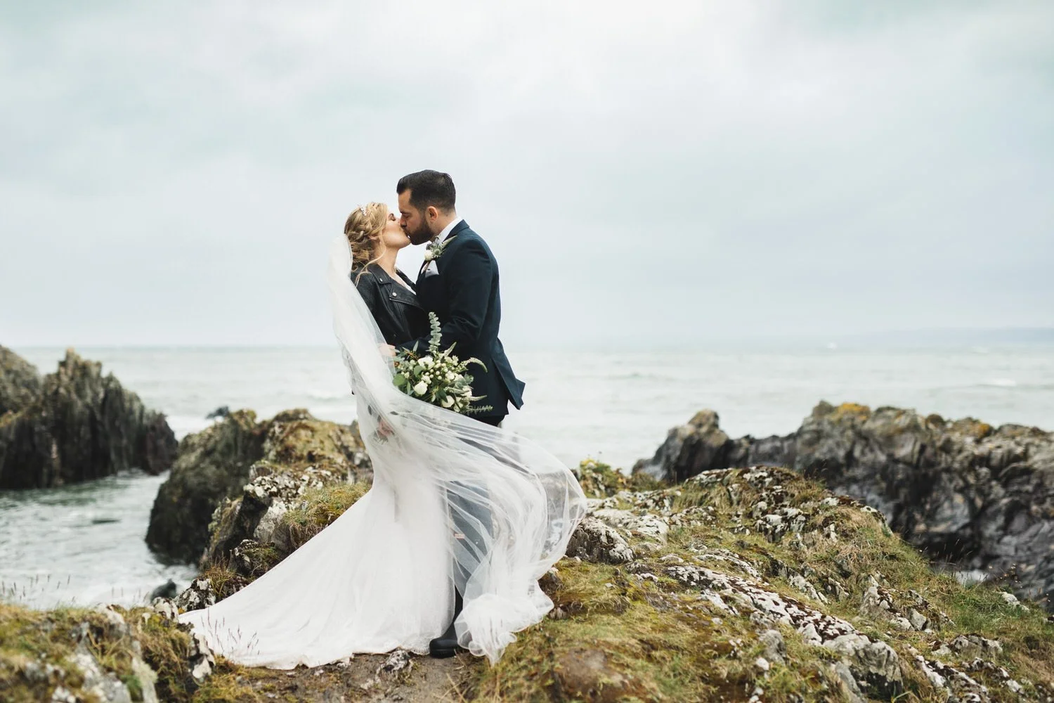 A bride and groom kissing on a rocky beach, with the bride holding a bouquet and wearing a long veil and white dress, and the groom in a dark suit against a cloudy sky and ocean background.