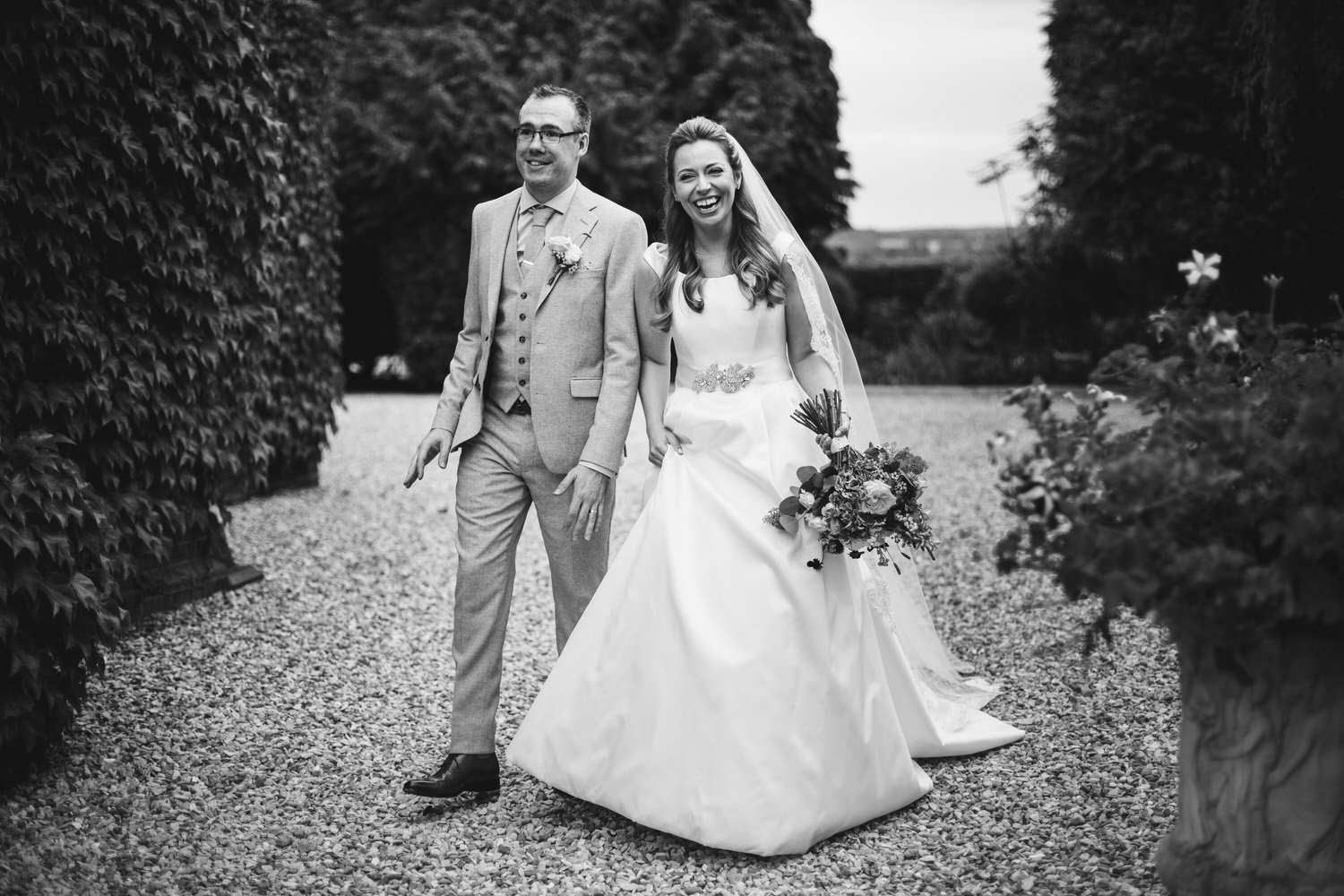 Black and white photo of a bride and groom walking outdoors, smiling, with greenery surrounding them. The bride is holding a bouquet, wearing a wedding dress and veil. The groom is dressed in a light-colored suit.