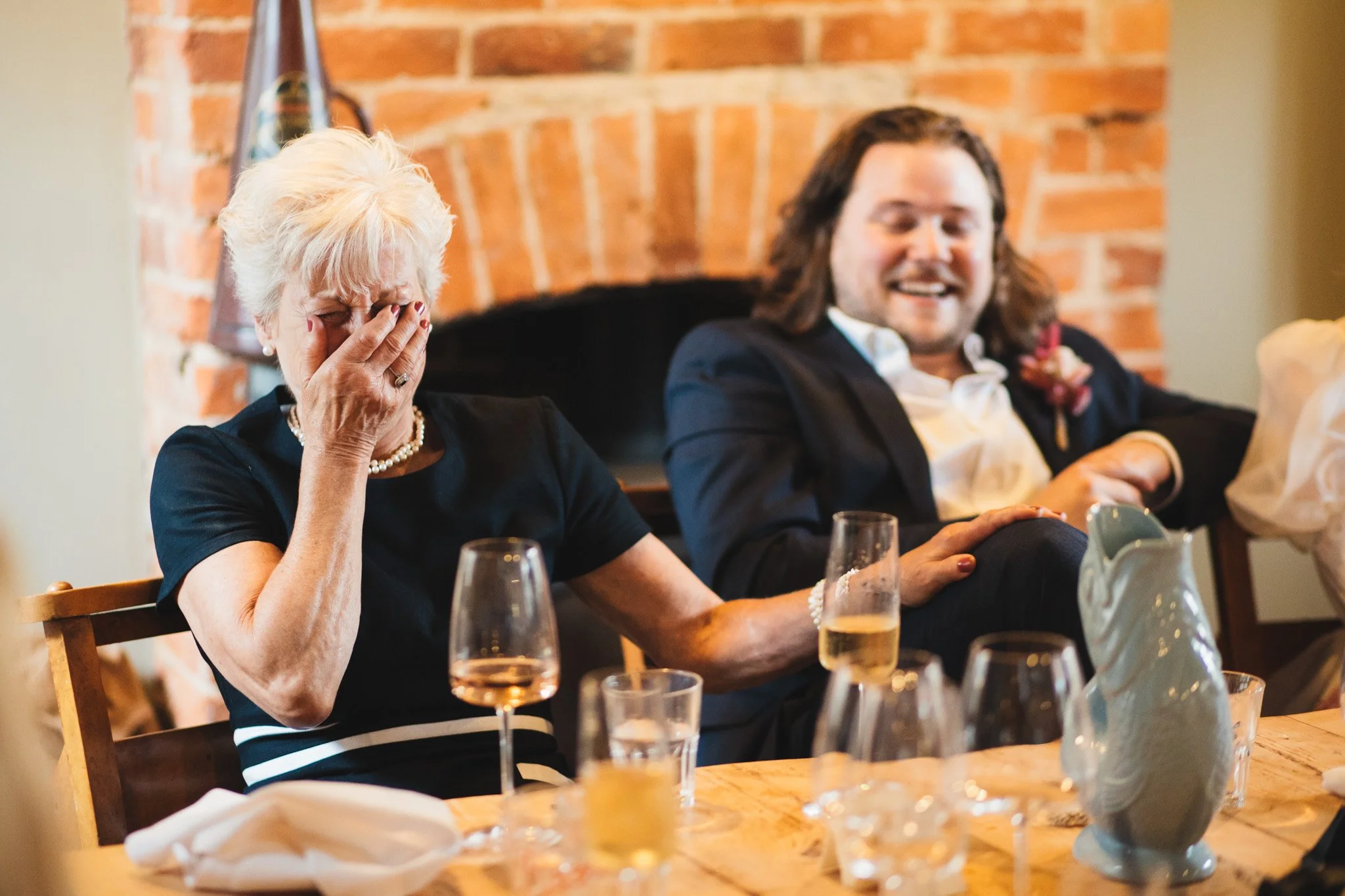 The groom's mother holds her hand to her face as she laughs at a joke in the wedding speeches.
