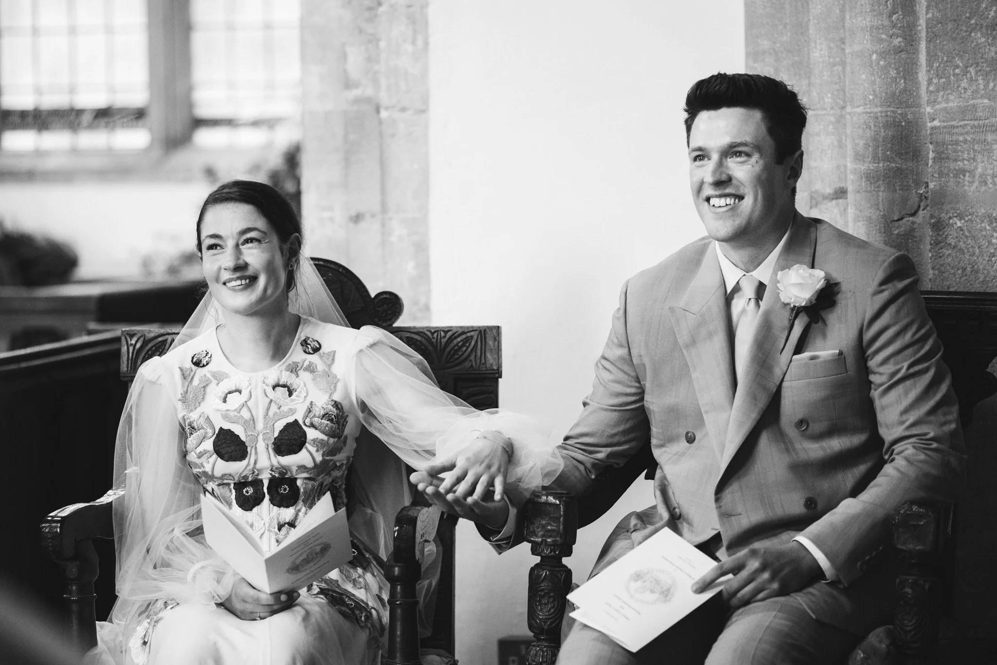 The bride and groom sit down and hold hands during their church ceremony. They look up and smile at the vicar.