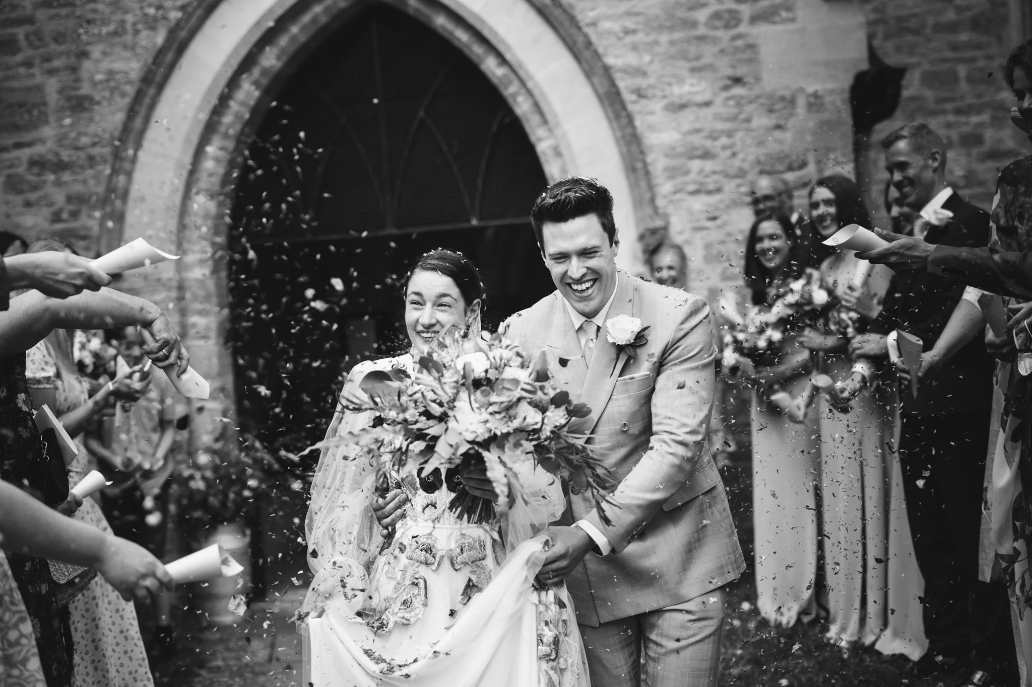 A black and white confetti photo. The  guests smile in the background as the couple laugh walking through falling dried petals.