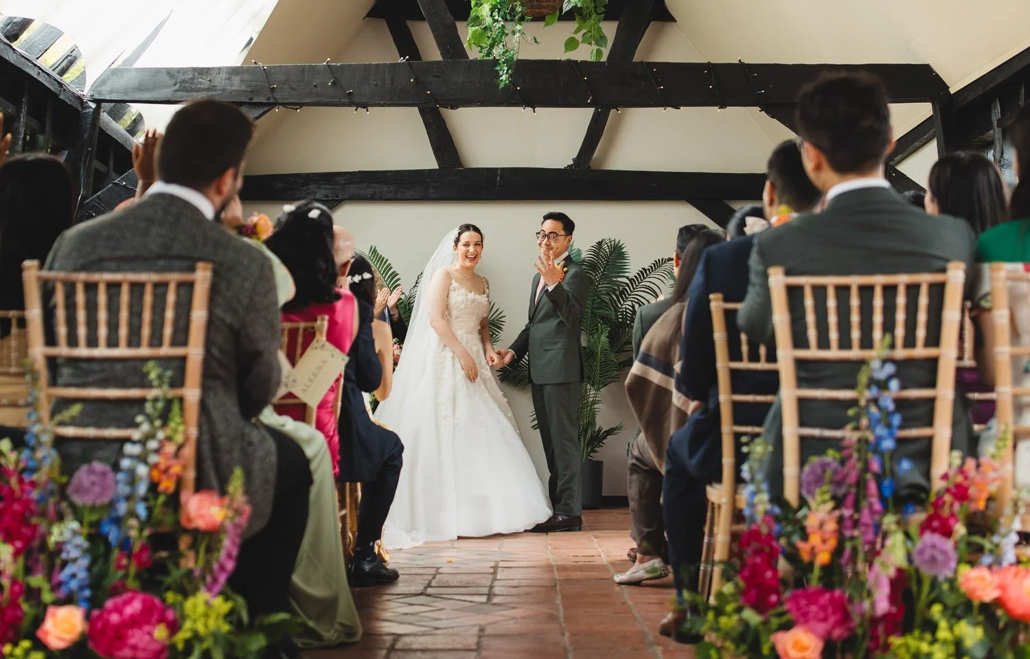 In the Old Dairy at South Farm, a bride and groom turn to their guests and laugh during the ceremony. The groom holds his hand up to them to show off his wedding ring.