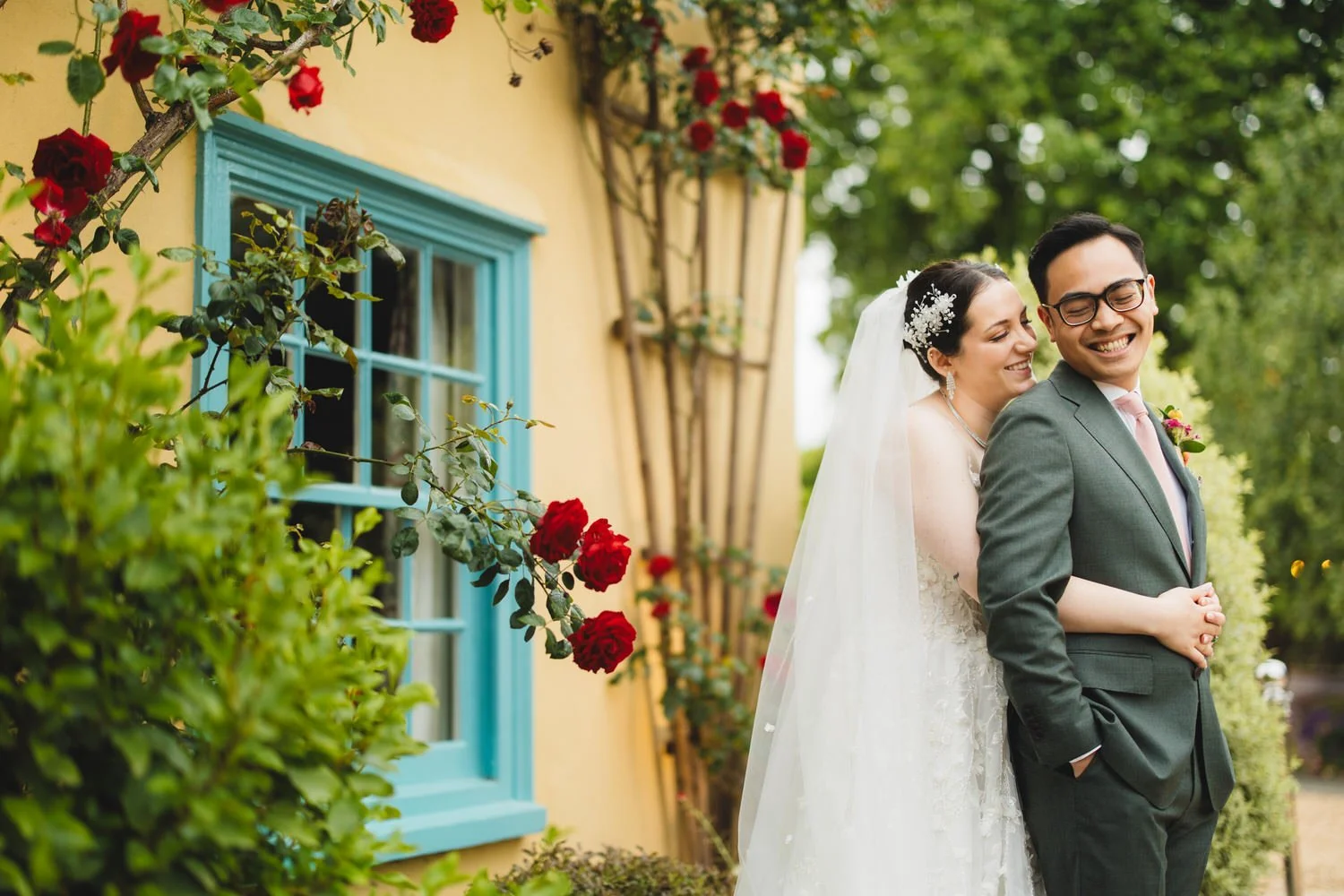 A bride hugs her groom from behind as they smile at each other. The South Farm farmhouse is in the backdrop, with a blue window and red roses.