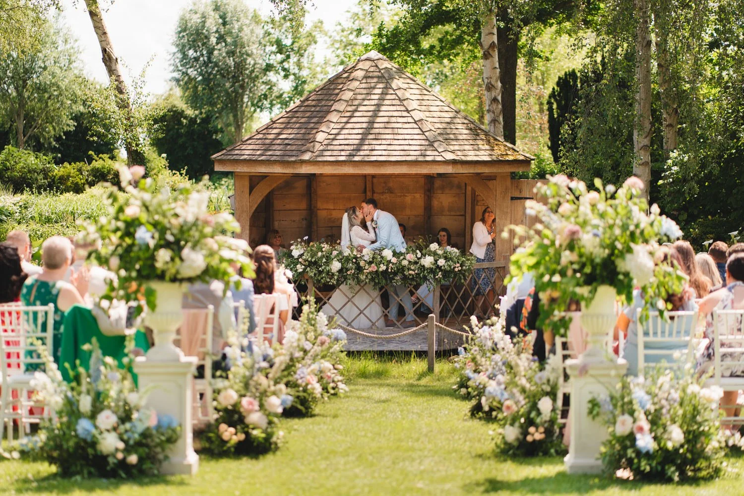 In the South Farm Summer House,  a newlywed couple kiss at the end of their wedding ceremony. The sun shines down as guests applaud.