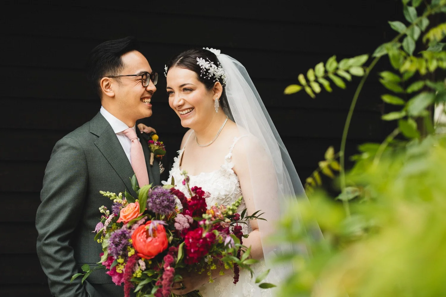A bride and groom laugh on their wedding day, with the bride holding a vibrant bouquet of flowers, and both dressed in wedding attire against a black background with some greenery in the foreground.