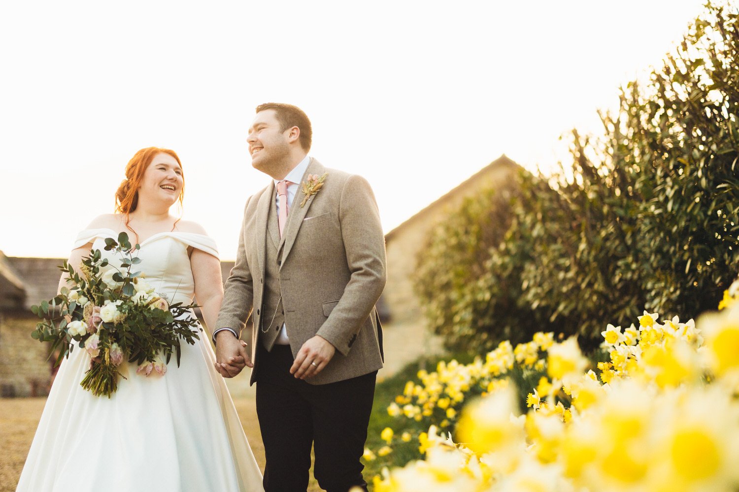 A bride and groom holding hands, smiling, walking outdoors during their wedding at Huntsmill Farm, with yellow flowers and greenery in the background, illuminated by warm sunlight.