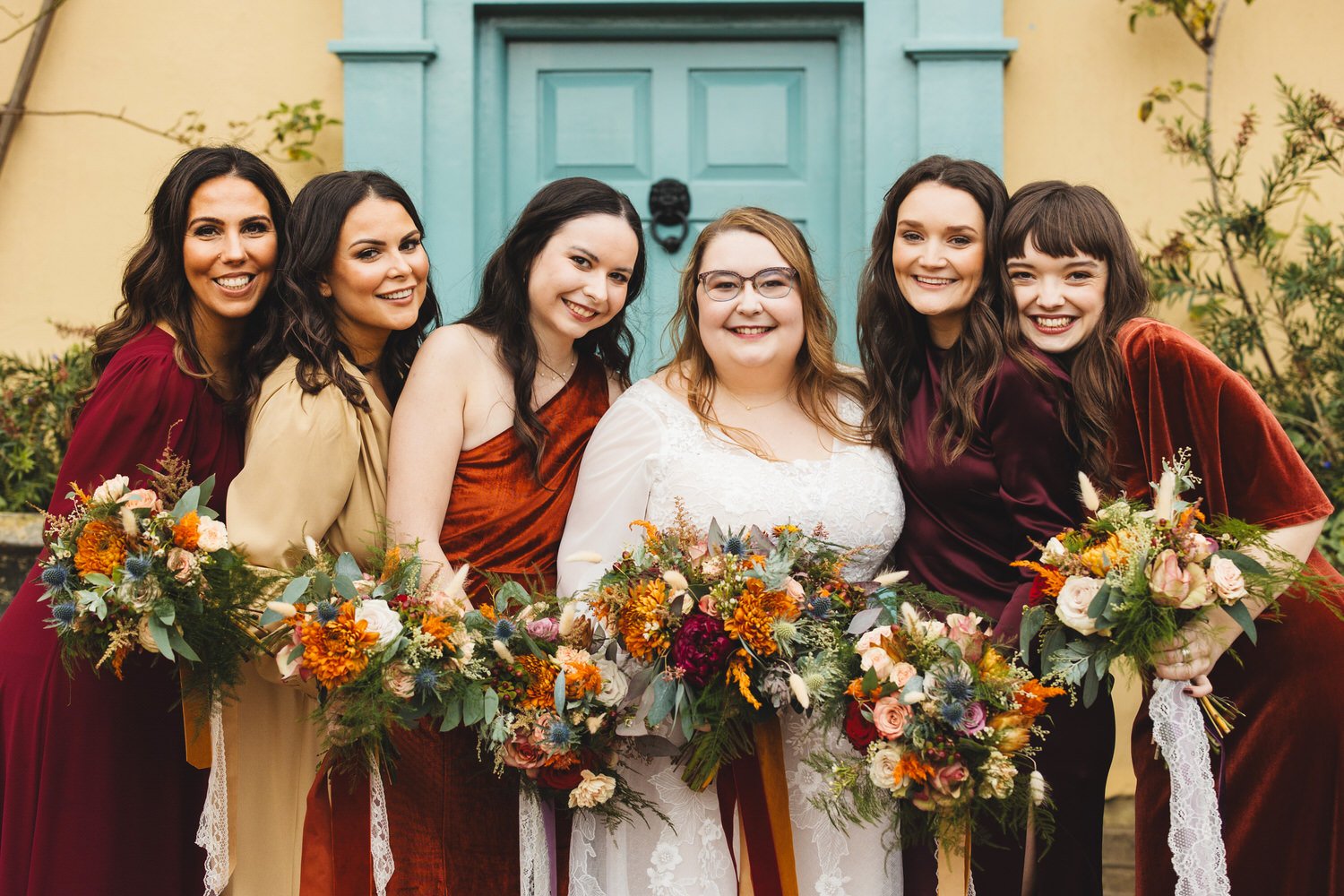 The bride and her bridesmaids cosy up together for a relaxed group photo. They all wear warm autumnal colours and carry bouquets tied with silk ribbons.