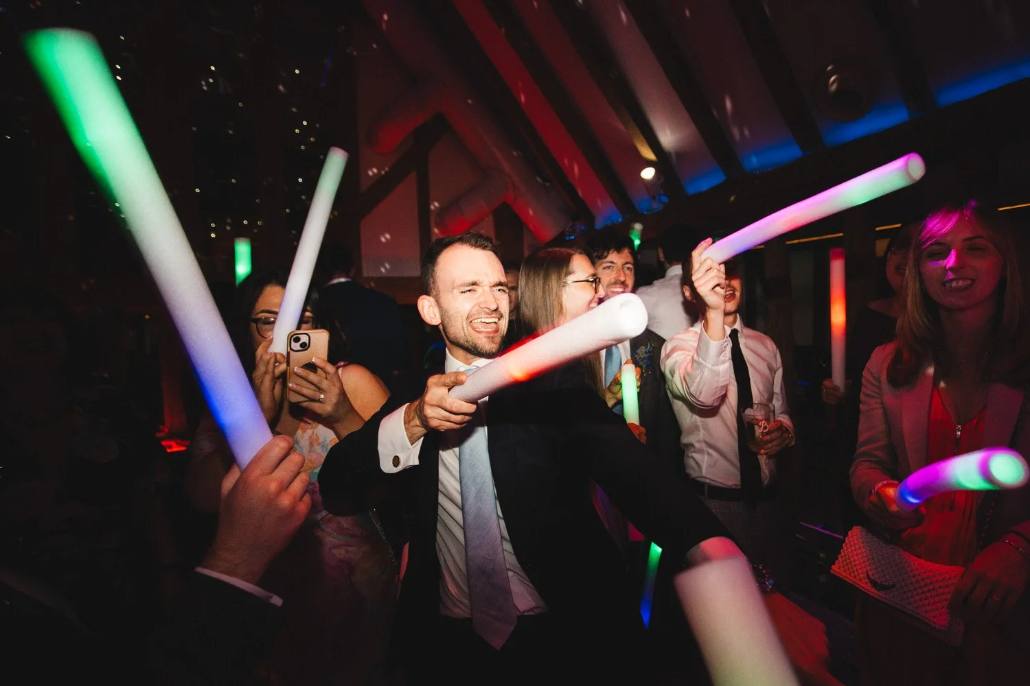 Guests sing as they dance to the wedding disco. They hold foam glowsticks and point them in the air.