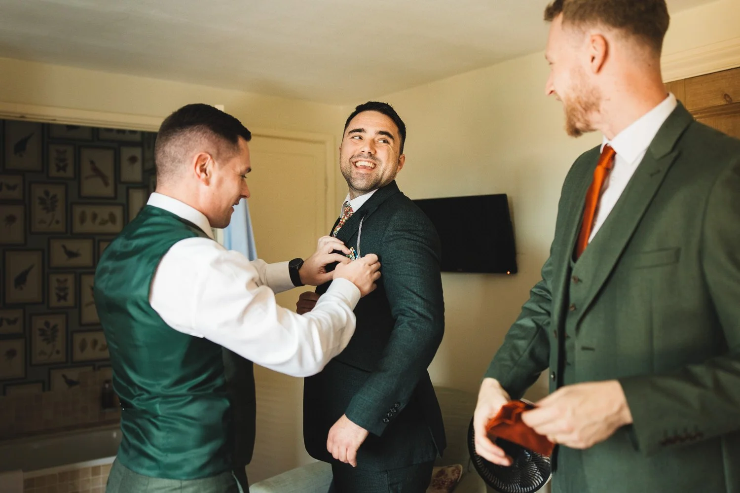 In the Parrot Room at South Farm, a groom in a dark green suit jacket laughs at one of his groomsmen as his best man arranges his pocket square.