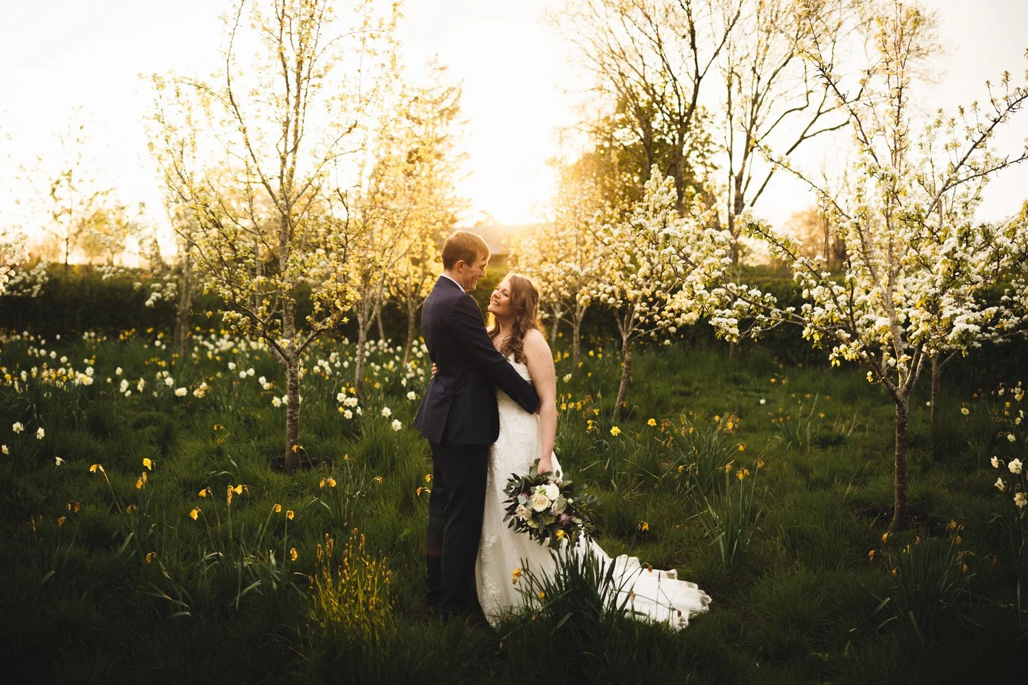 The sun sets behind an embracing couple in the Orchard at South Farm. The apple trees are covered in white blossom.