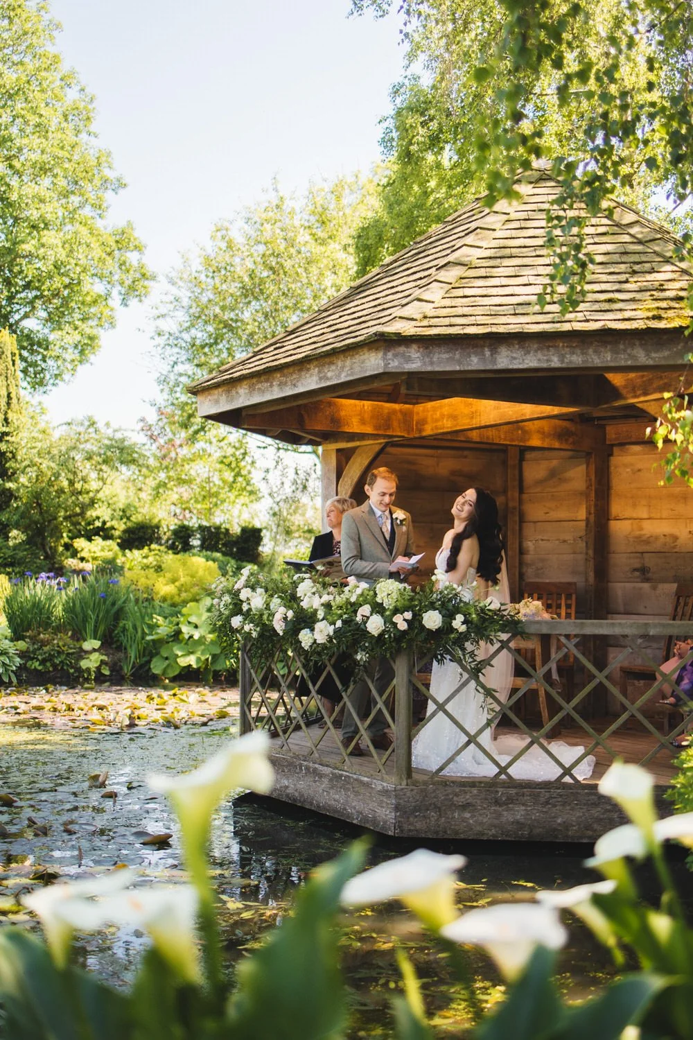 During their wedding ceremony in the Summer House at South Farm, the groom reads his vows to the bride as she laughs. The Summer House sits on a lily pond.