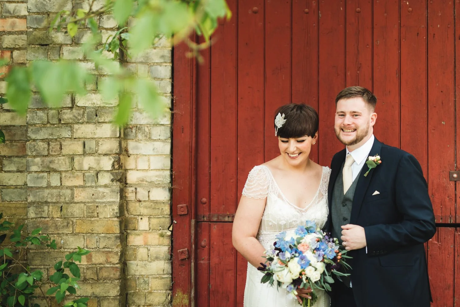 A happy bride and groom standing together in front of a red wooden farm door, smiling and holding a colourful bouquet of flowers.