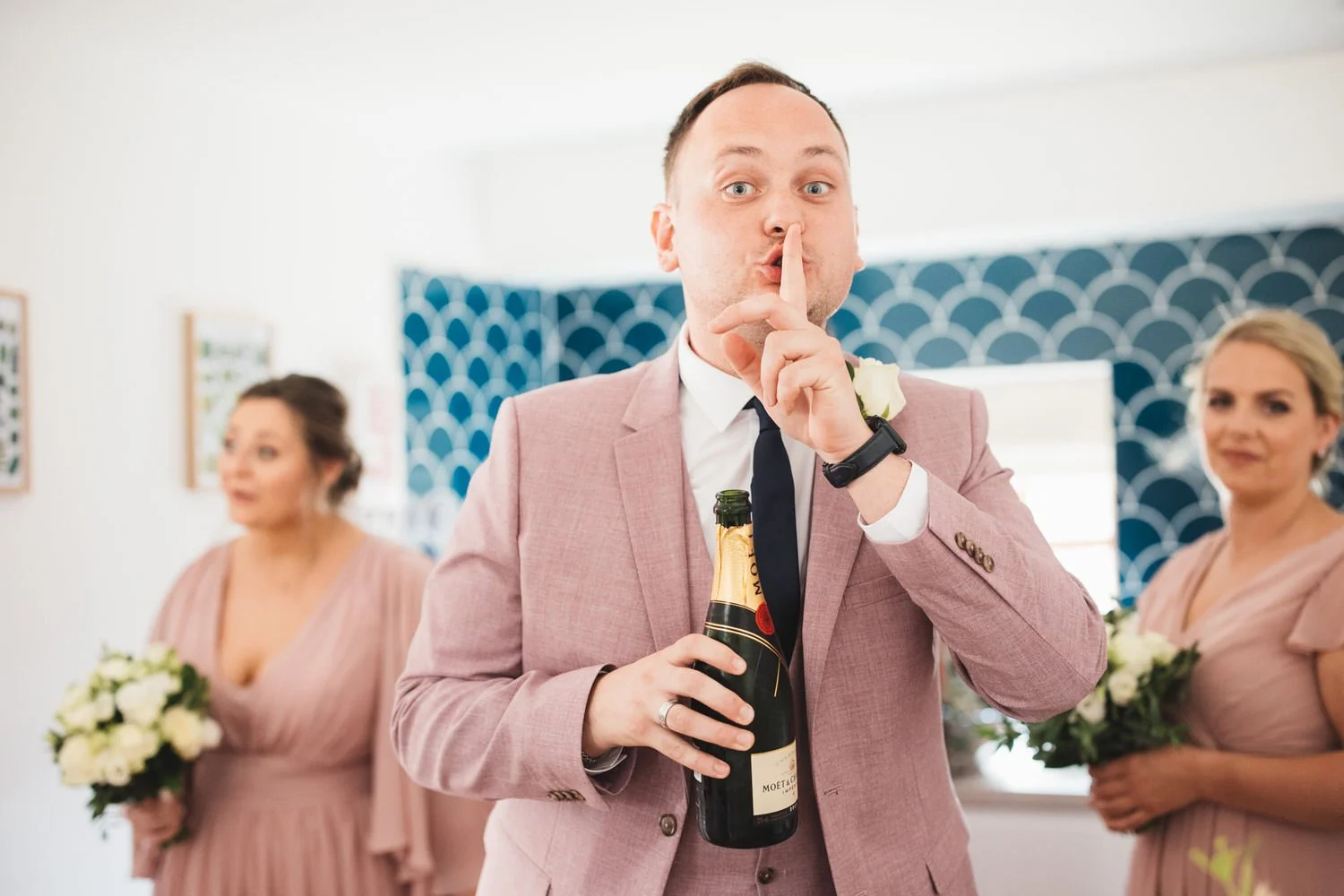 One of the bride's friends holds a finger to his lips in a "ssh" pose as he holds an open bottle of Moët champagne on the wedding morning. He wears a dusty pink suit and the bridesmaids are in the background.