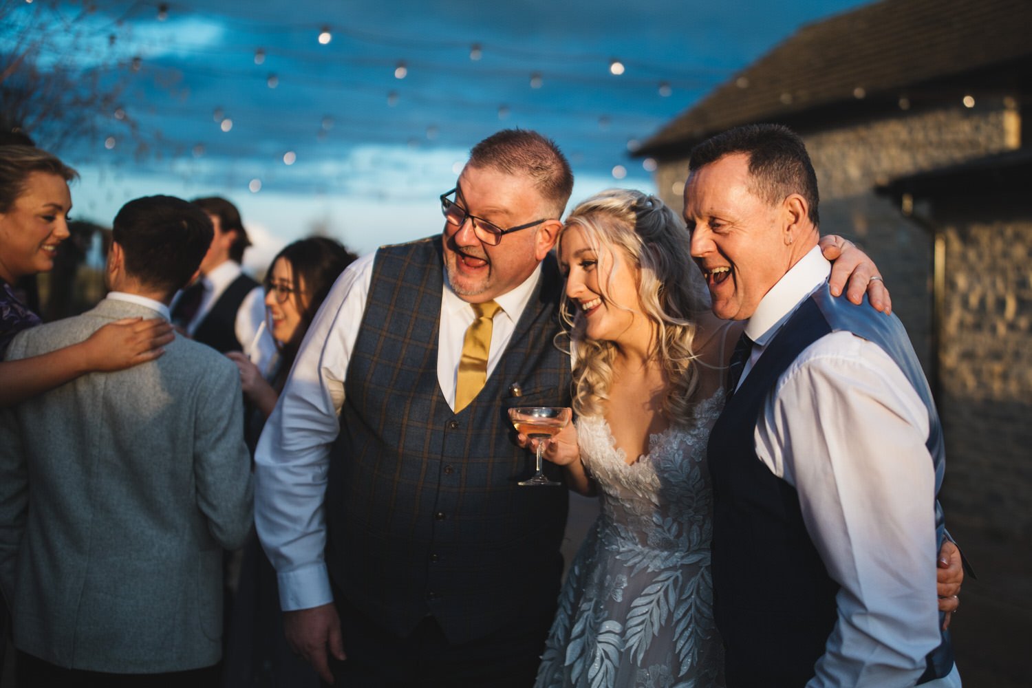 The bride hugs two wedding guests in the evening sun as a friend takes a photo of them on their phone.