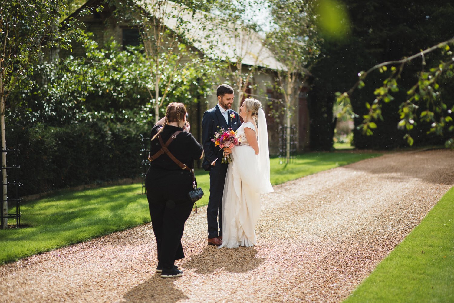 A bride and groom stand on a gravel path during their wedding ceremony outdoors, smiling at each other, with Cat Lane taking a photograph of them, surrounded by green trees and grass.