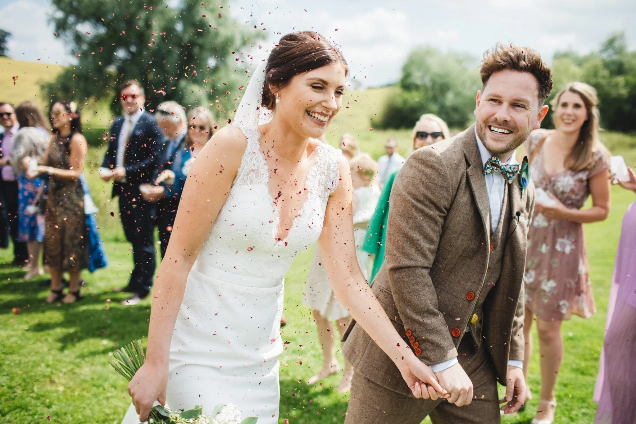 A joyful confetti moment, the bride and groom smile as guests throw red petal confetti.