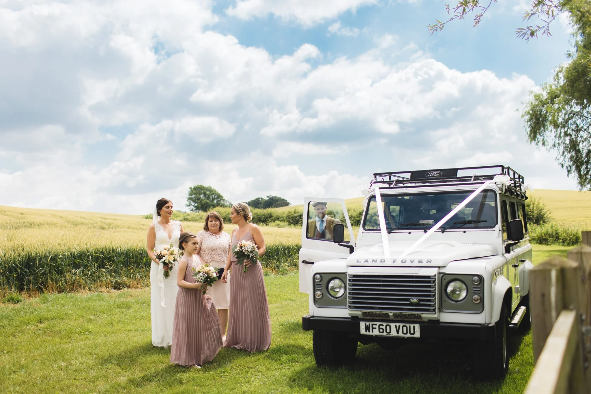 In front of a countryside landscape, the bride and her bridesmaids leave the Land Rover wedding car before the ceremony. There are Bedfordshire wheat fields and blue skies behind them.