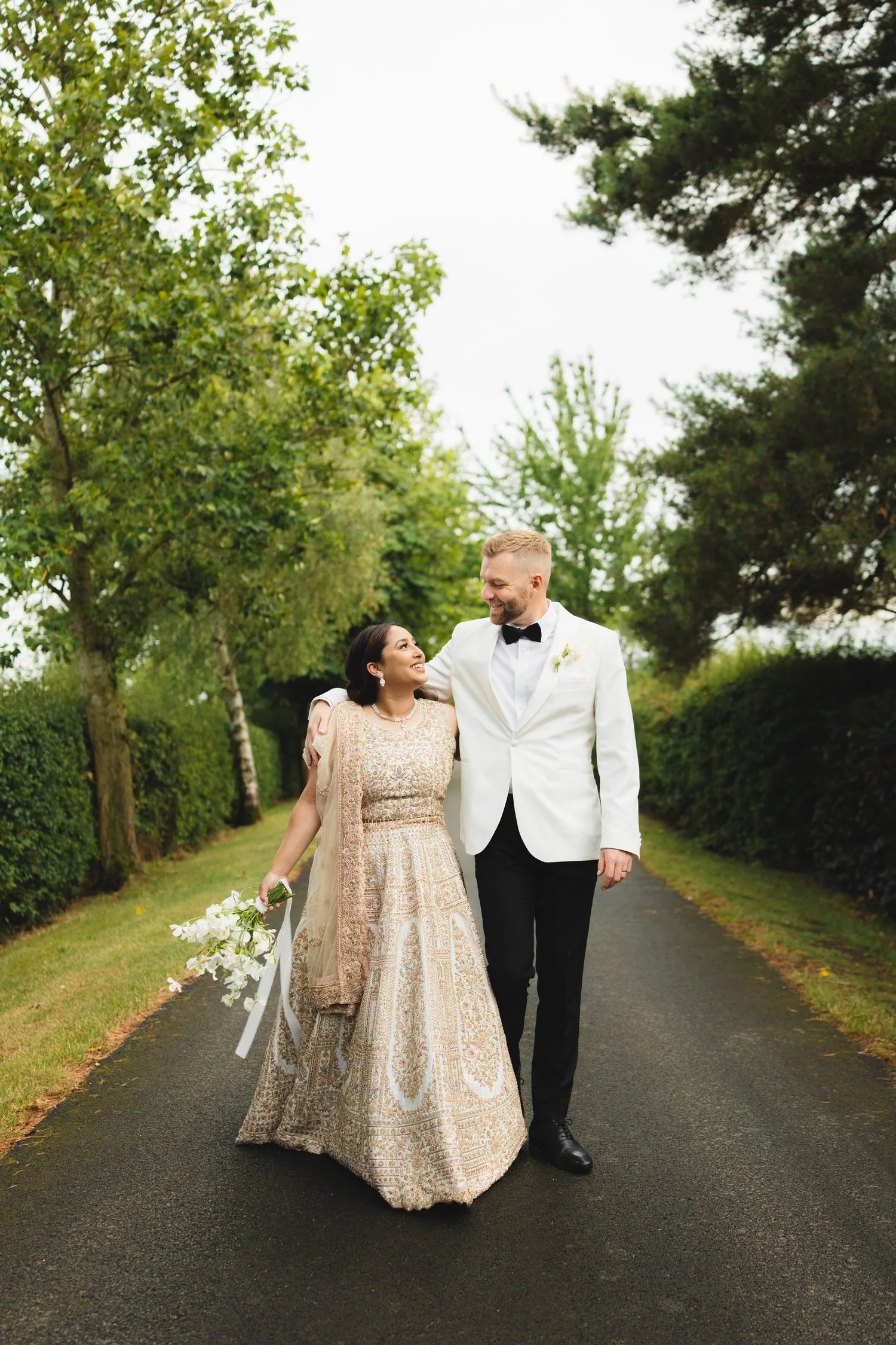 A groom in a white tuxedo puts his arm around his bride in a gold embroidered bridal sari. They walk down a countryside road with greenery behind them and smile at each other.