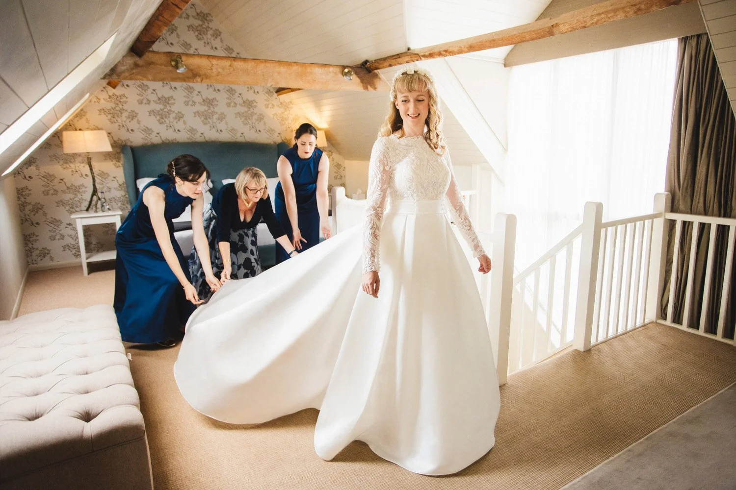 In the Coach House Suite at South Farm, a bride smiles as her mother and bridesmaids fluff out the train of her wedding dress.