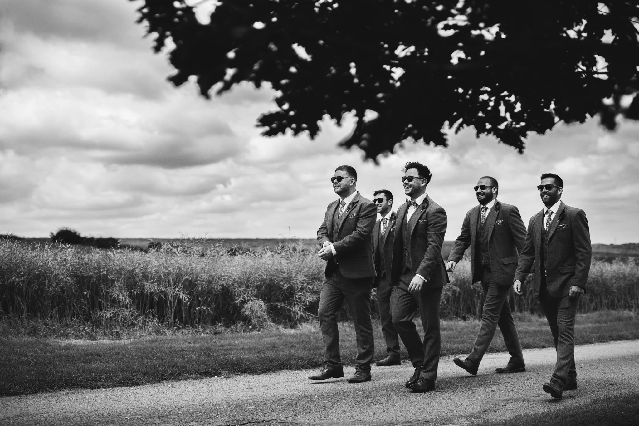 A black and white candid photo of the groom and the groomsmen walking to the wedding. They wear tweed suits and sunglasses.
