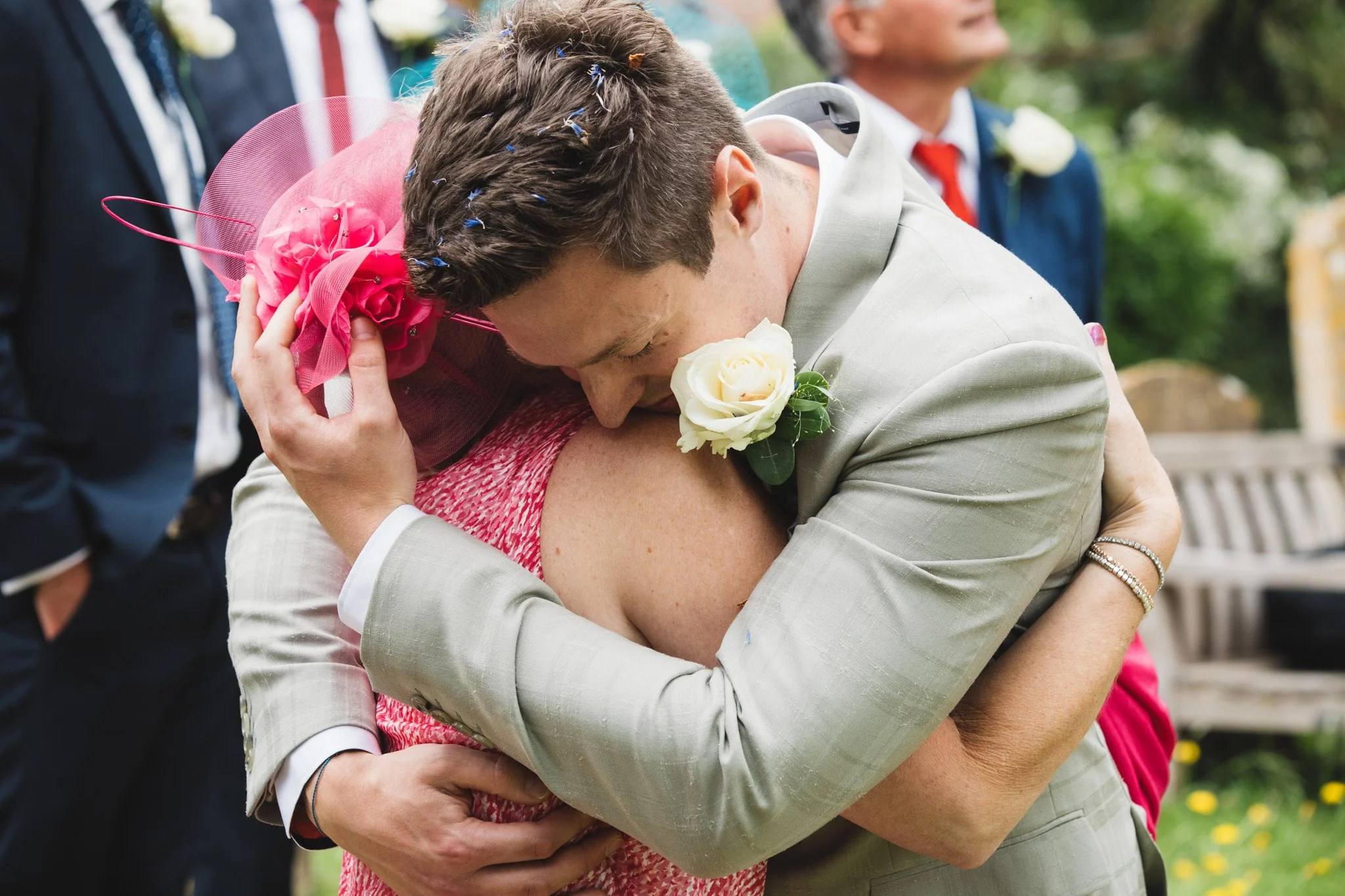 The groom tightly hugs his mother after the wedding ceremony. She wears pink and he has confetti in his hair.