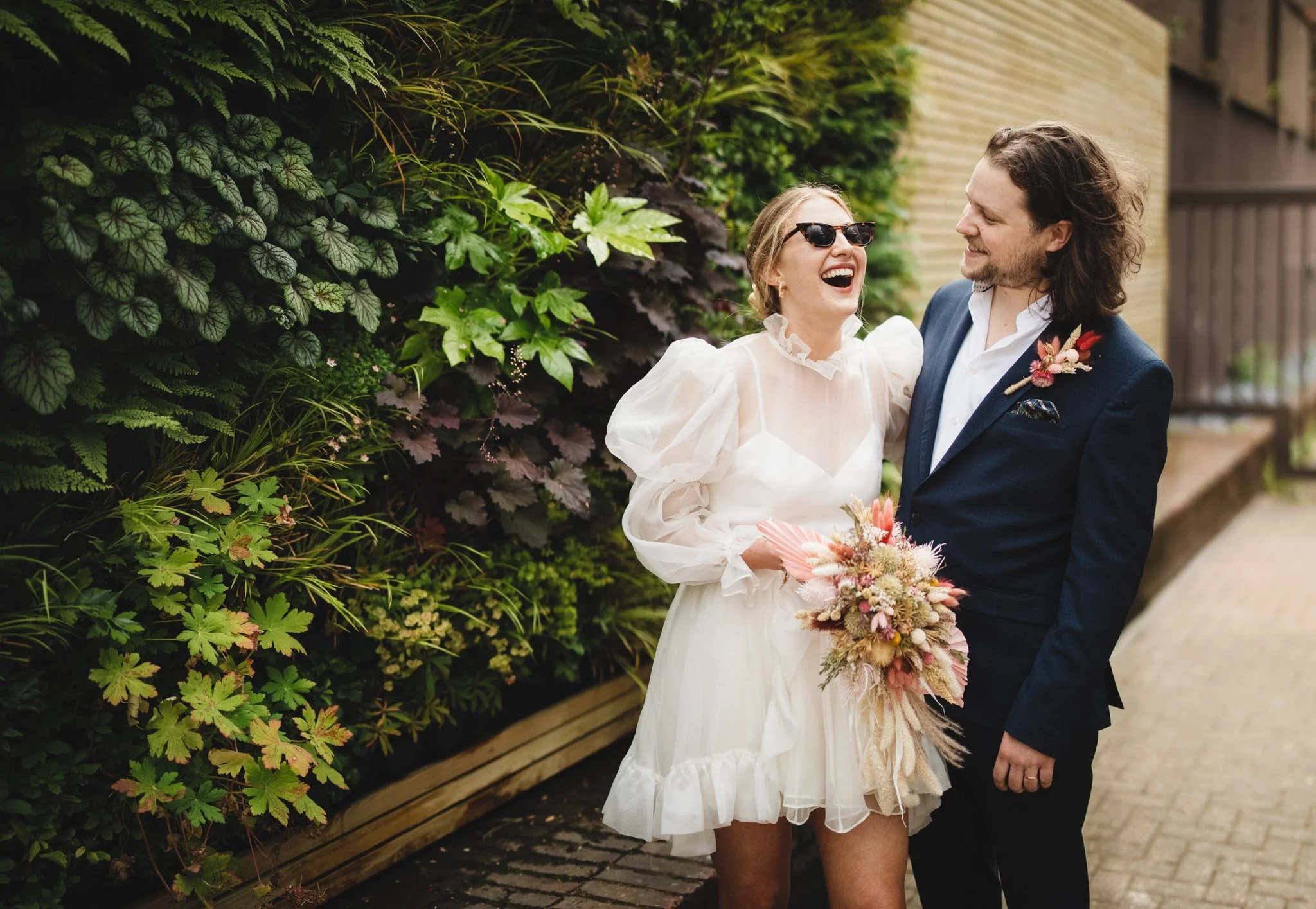During their micro wedding in Bedford, a couple hug and laugh together in front of a wall of plants. The bride wears sunglasses and looks upward laughing.