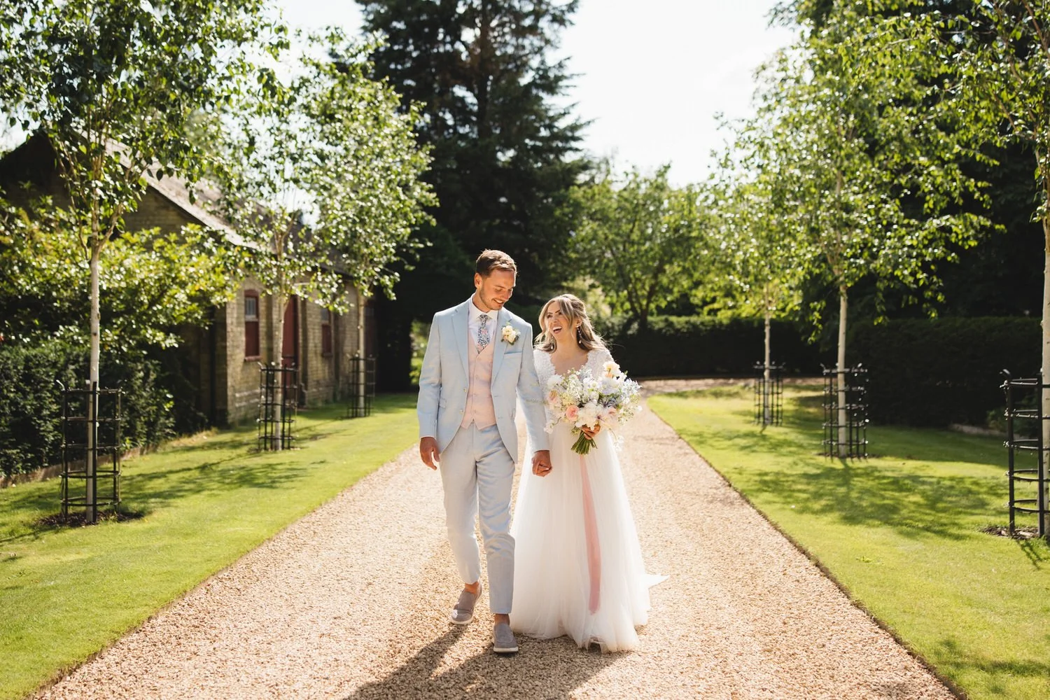 On a hot summer day, a couple walk down the South Farm driveway laughing and chatting. The groom wears a pastel blue suit with pink waistcoat, and the bride wears a tulle dress and carries a bouquet of pastel flowers.