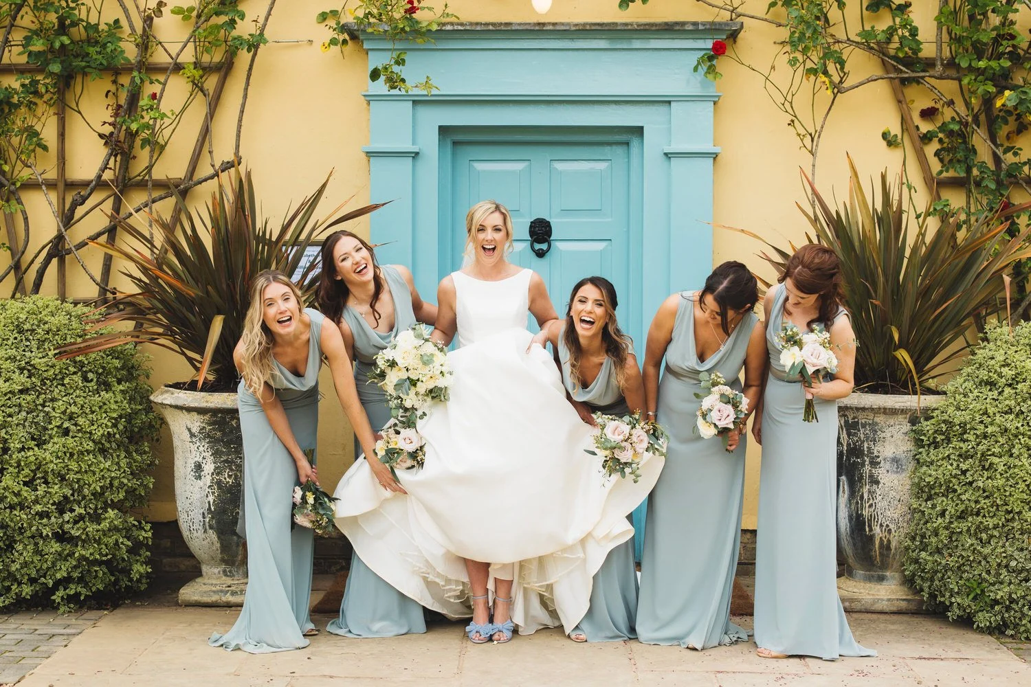 The bride laughs and points to her feet as her bridesmaids lift up her dress to show off her wedding shoes, pale blue to match the South Farm door.