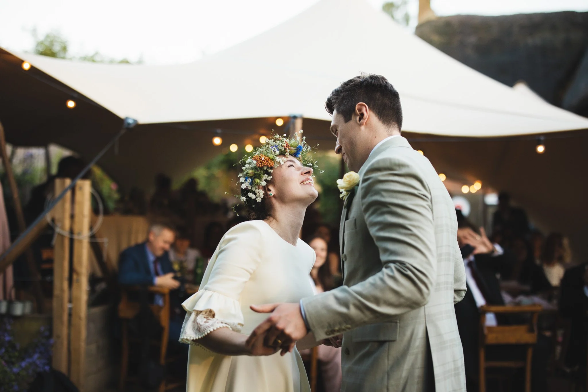 The bride and groom hold hands as they have their first dance outside. Guests look on in the background.