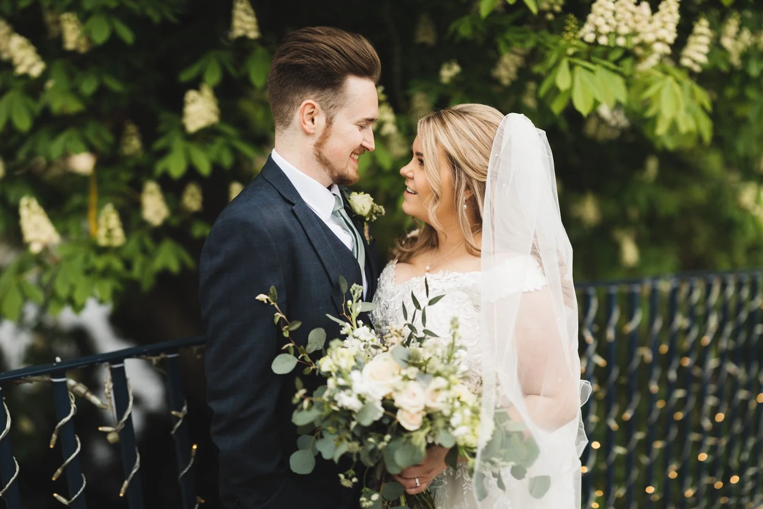 A bride and groom standing close together outdoors, smiling and looking into each other's eyes, with the bride holding a bouquet of white and green flowers on the bridge at Bassmead Manor Barns.