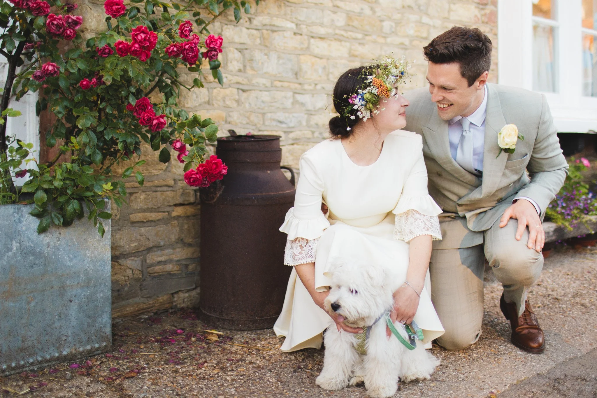 The bride and groom share a cute look as they hug their little fluffy dog with pink roses in the background.