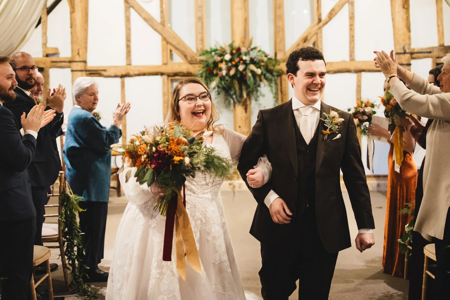 A couple cheer and smile as they walk back down the aisle in South Farm's Tudor Barn. The beams of the barn and a floral installation are behind them and their guests are applauding.