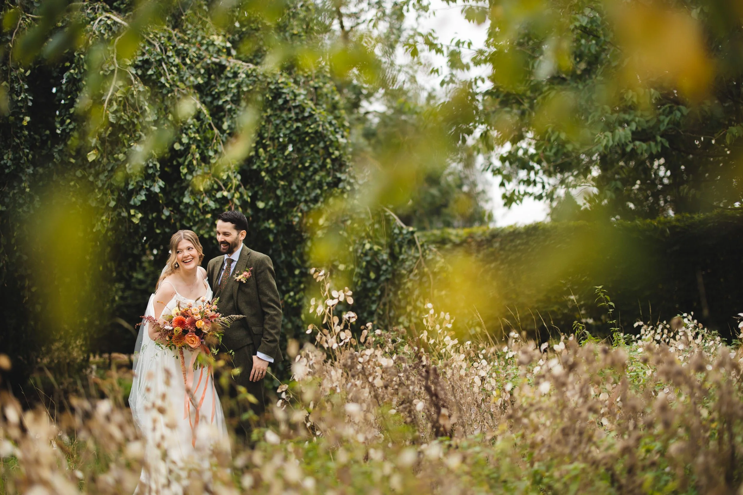 A bride and groom smiling and holding a bouquet of colourful flowers, surrounded by autumn greenery and flowers at the Barn at Redcoats in Hertfordshire.