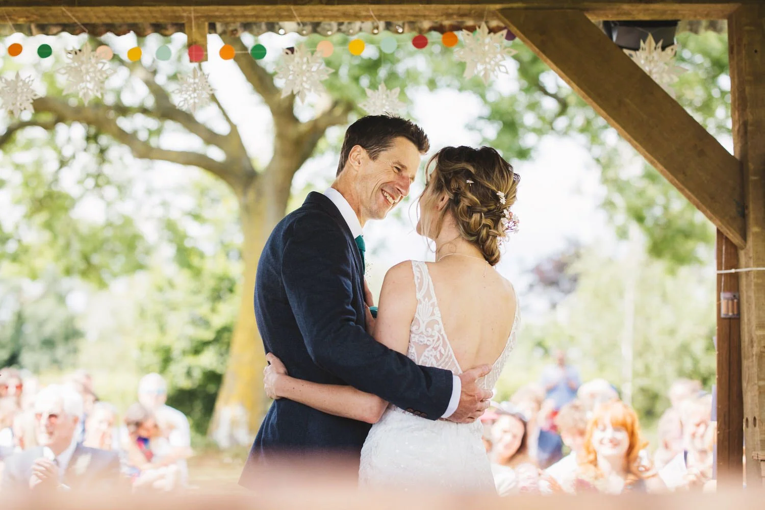 A bride and groom embrace at the end of their outdoor wedding ceremony at Huntsmill Farm in Buckinghamshire. Their guests smile in the background and colourful pom poms hang from the shed ceiling.