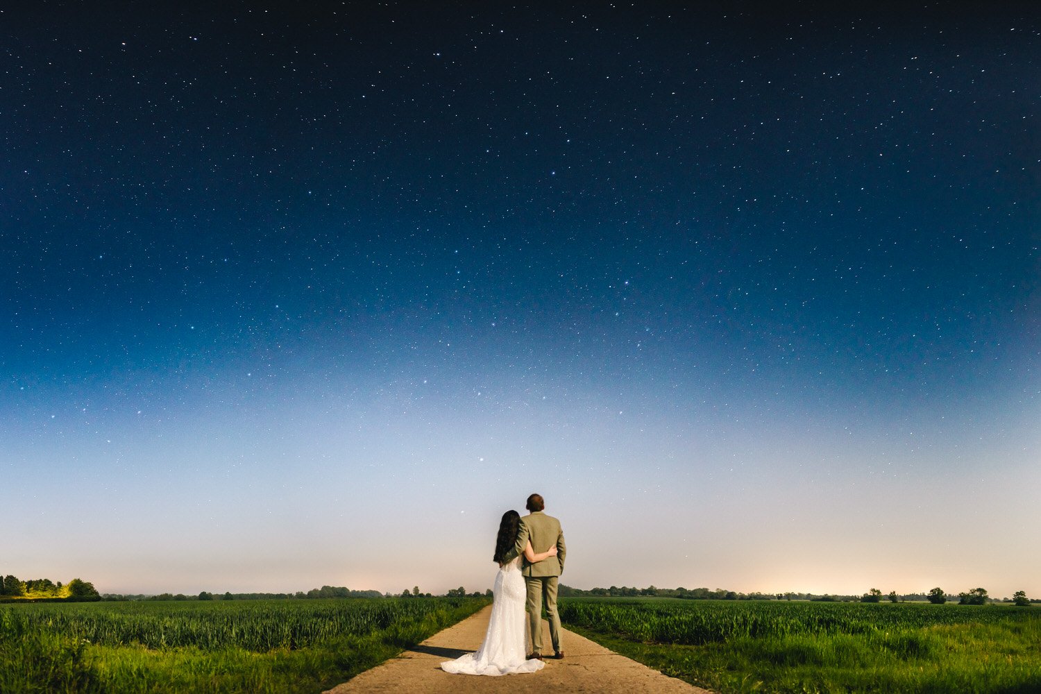 A couple in wedding attire stand on a dirt path in a field at night, gazing up at a brilliant blue starry sky over South Farm in Hertfordshire.