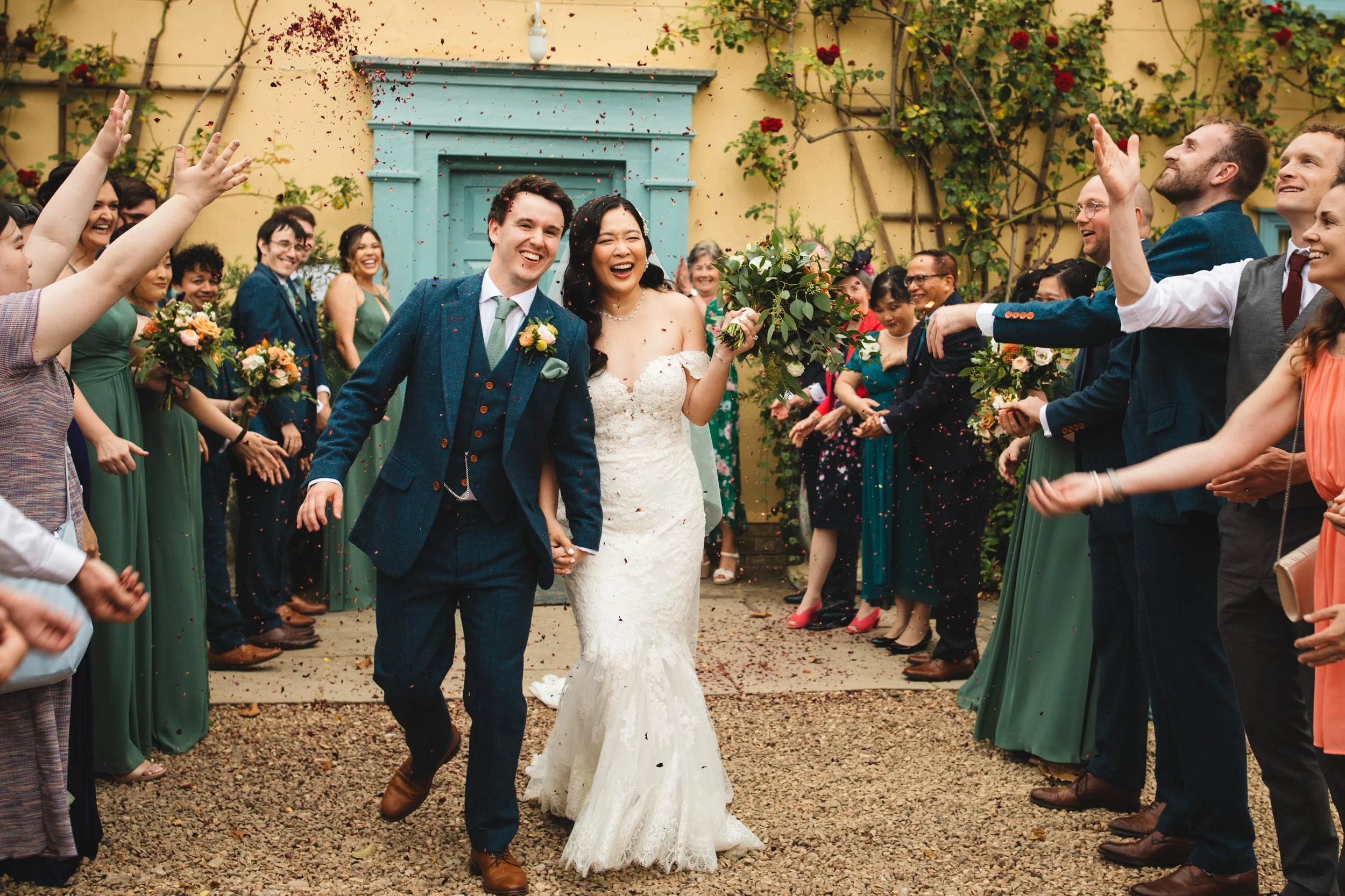A couple joyfully run forward as smiling guests throw petal confetti over them with the South Farm blue doors in the background. The groom is wearing a blue tweed suit, and the bride is wearing a sleeveless lace dress.