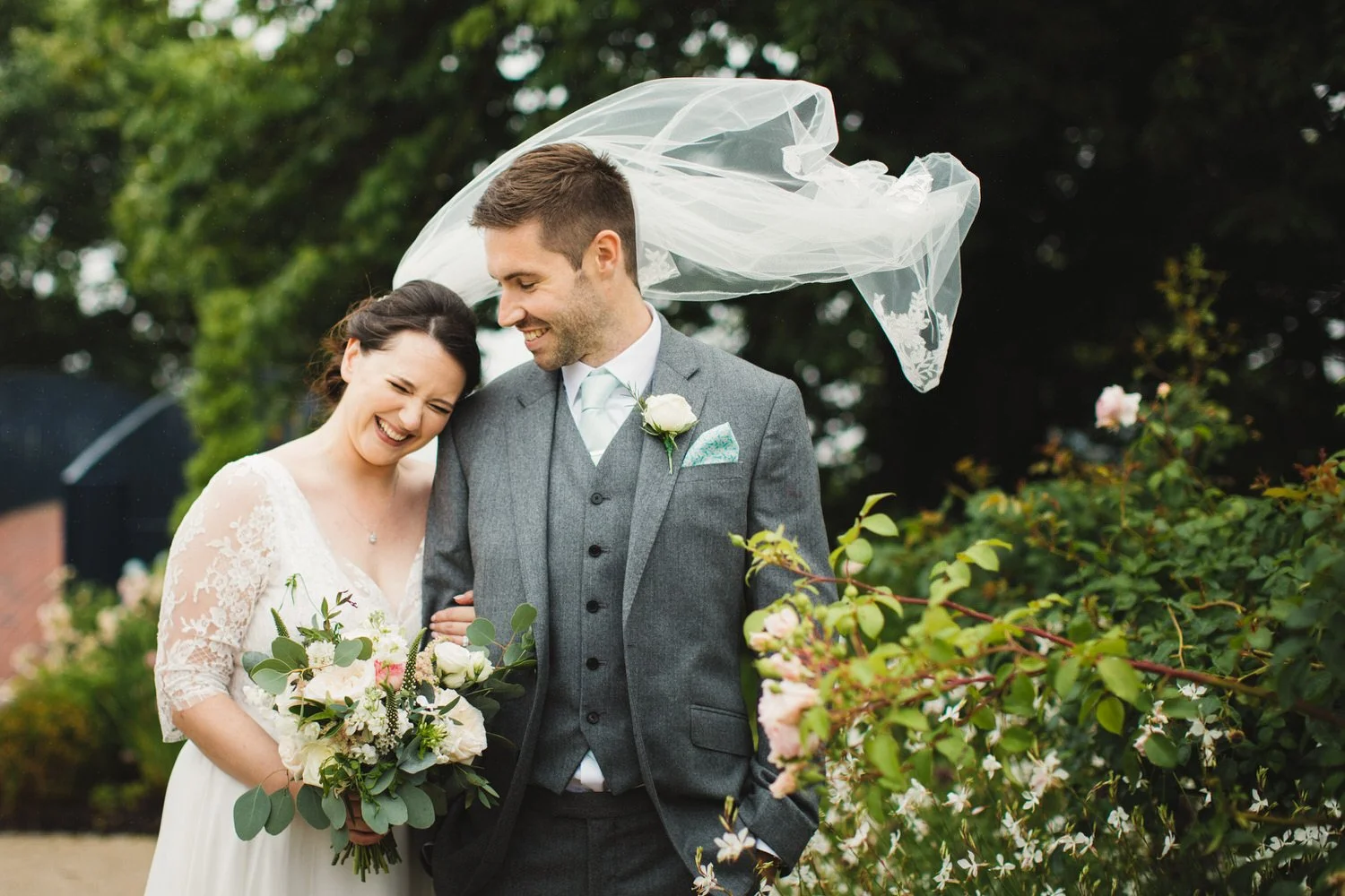 A newlywed couple smiling and laughing outdoors, with the bride holding a bouquet of flowers and the groom in a gray suit with a boutonniere, in front of green shrubbery at Bassmead Manor Barns.