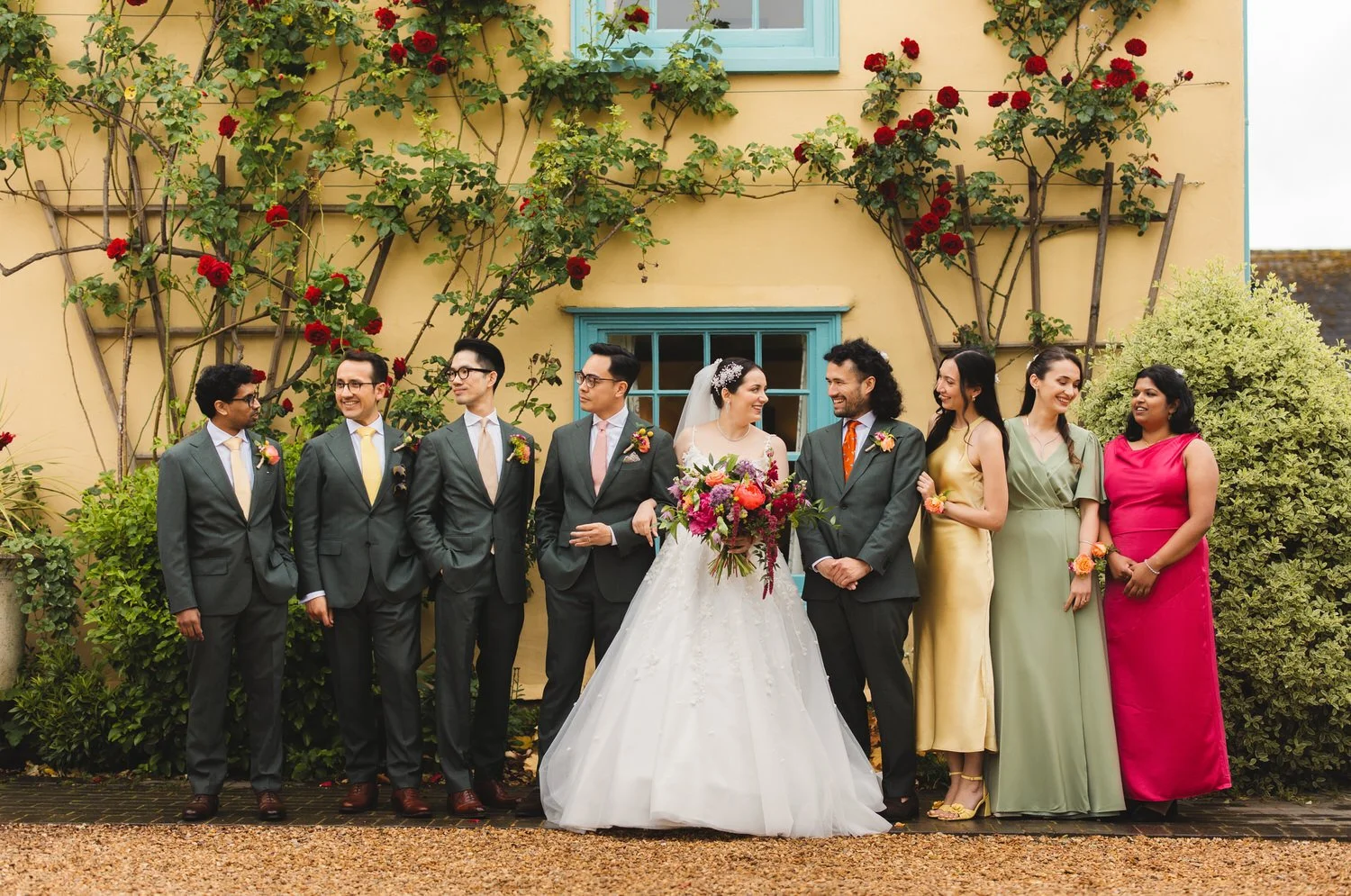 A couple and their friends stand for a group photo in front of the South Farm farmhouse and red roses. The bridesmaids are wearing brightly coloured dresses, and the groomsmen wear dark green suits. They look at each other and chat, smiling.