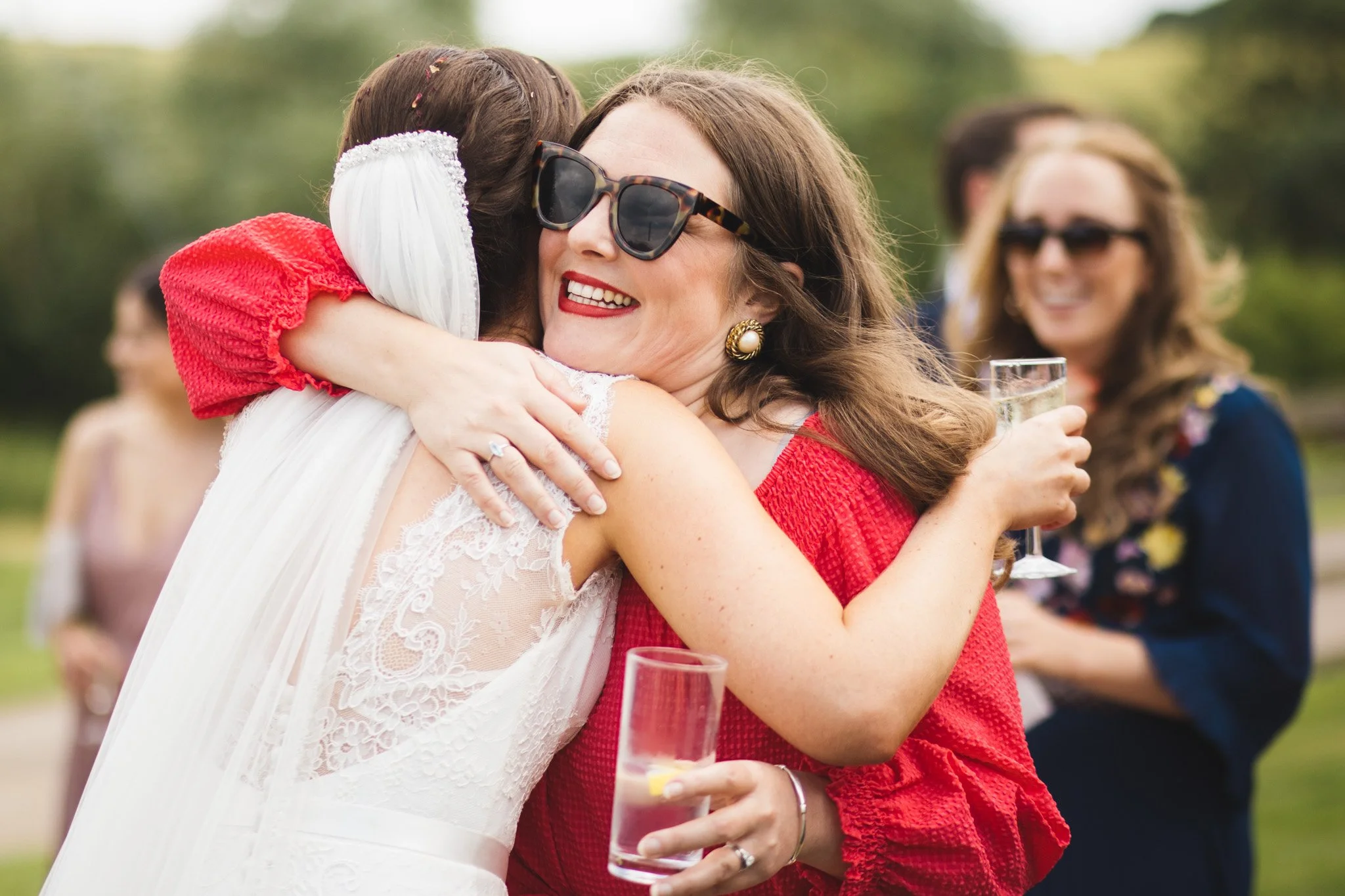 A wedding guest hugs the bride tightly as she congratulates her. The wedding guest is in red and other guests smile in the background.