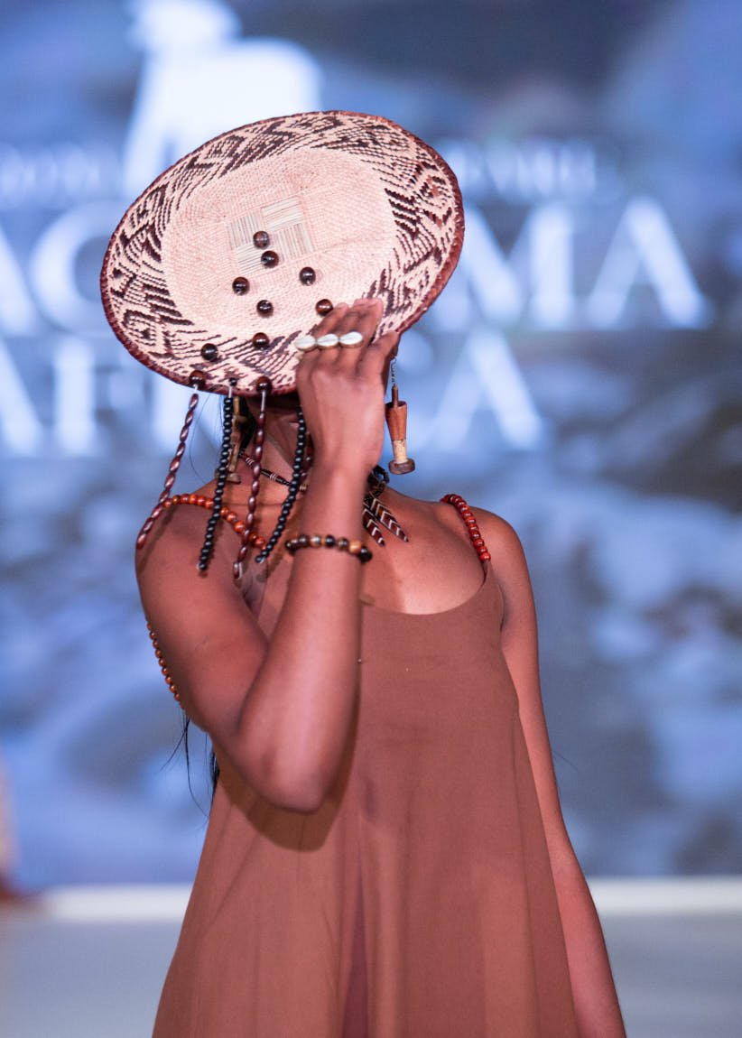 Runway model wearing a brown sleeveless dress and holding a circular patterned accessory in front of her face.