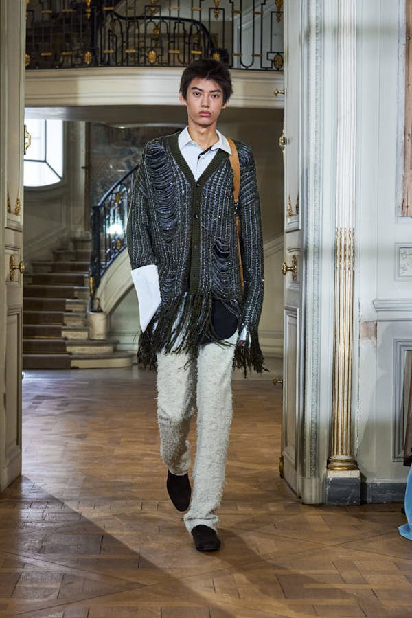 Model walks the Dior Men Summer 2027 runway in a textured grey coat, white shirt, black bow tie and wide black trousers inside the Musée Nissim de Camondo