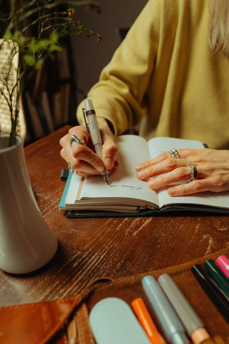 A person writes in an open notebook with a Kaweco pen at a wooden desk, surrounded by flowers, a leather pen case, and colourful writing instruments