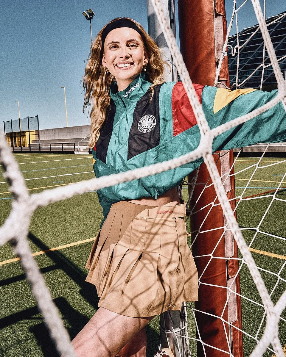 A model wears an adidas Originals Germany EQT track jacket and pleated skirt while standing inside a football goal on a sunny pitch