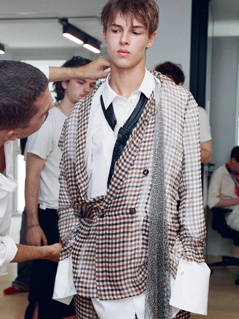 Model backstage at Dior Men Summer 2027 wearing a checked suit with extended white shirt cuffs and a loosely tied black scarf for LE MILE Magazine