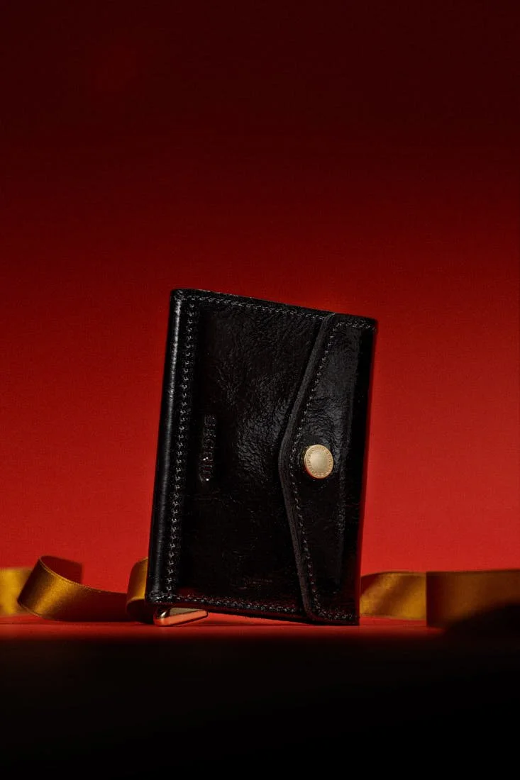 SECRID Premium Envelope Wallet+ Naplak in black with glossy leather, stitched envelope construction and gold-tone fastening, photographed against a deep red background for LE MILE Magazine.