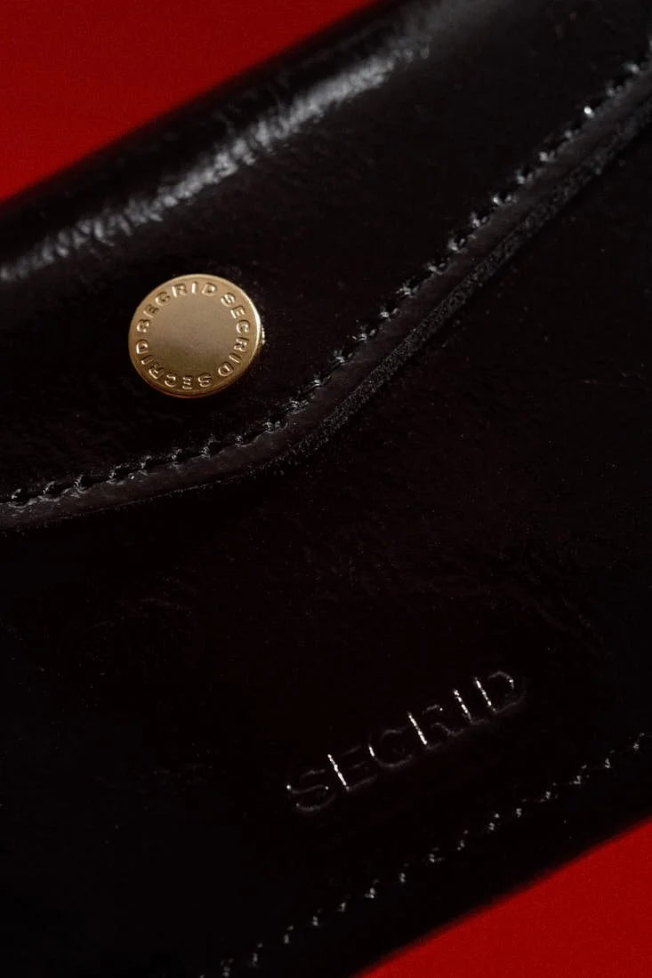 Close-up of the SECRID Premium Envelope Wallet+ Naplak in black, showing the glossy leather surface, stitching, embossed SECRID logo and gold-tone fastening detail, LE MILE Magazine.
